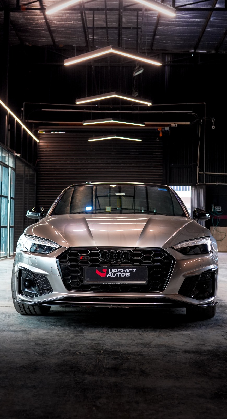 A silver Audi S5 Sportback head-on in the workshop, its black honeycomb grille behind the Upshift plate.