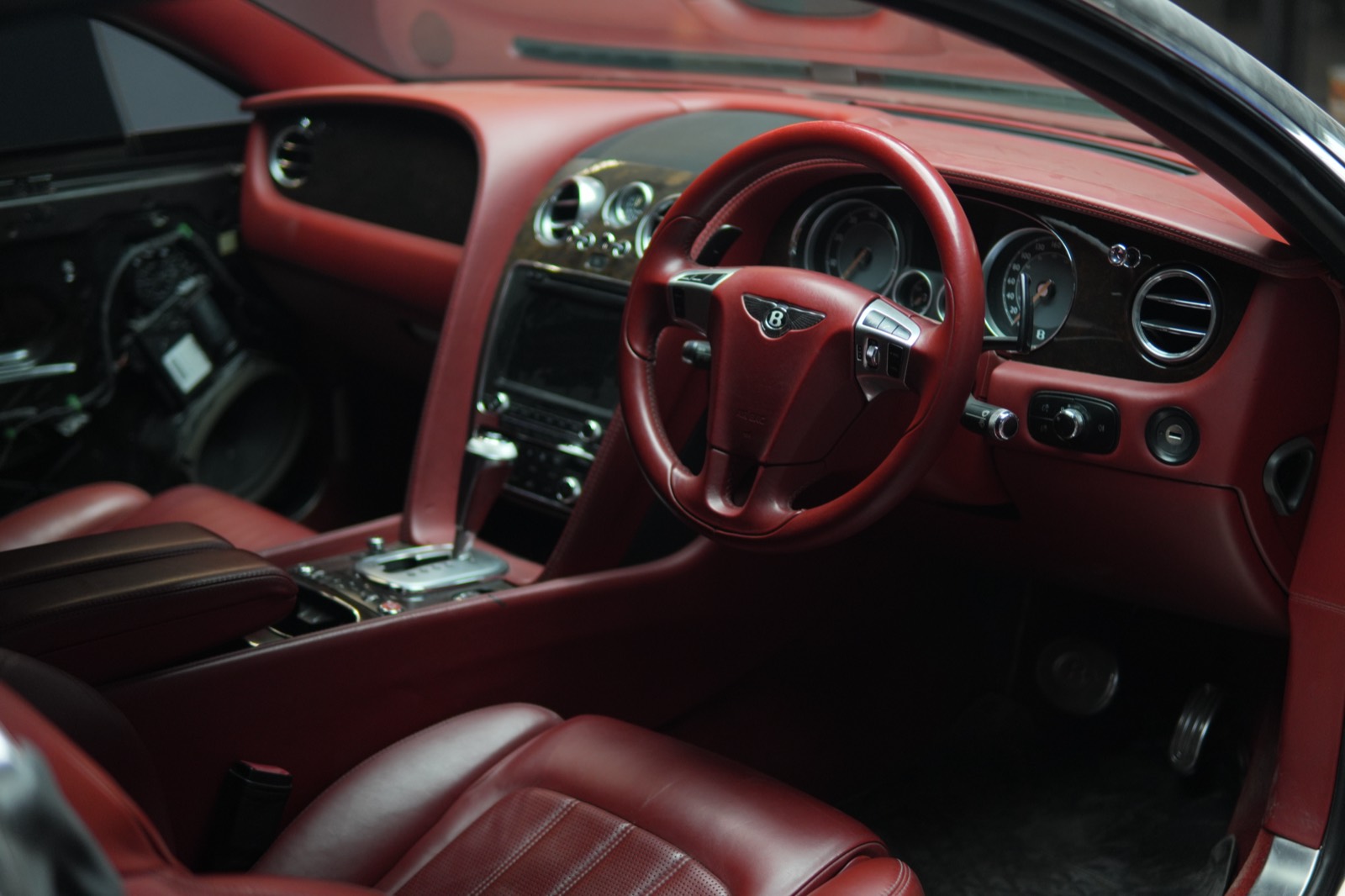The red leather cabin of a Bentley Continental seen from the driver's door, with dark wood veneer on the dash and the trim panel removed from the far door, its wiring exposed.