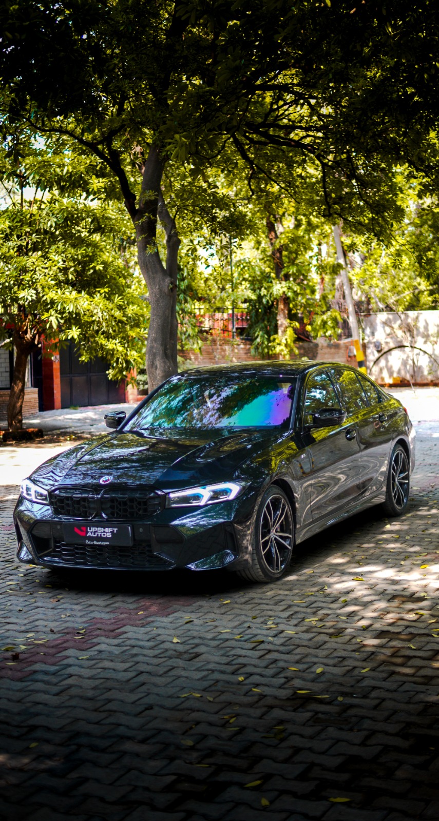 A black BMW 3 Series on a paved courtyard under trees, front three-quarter view.