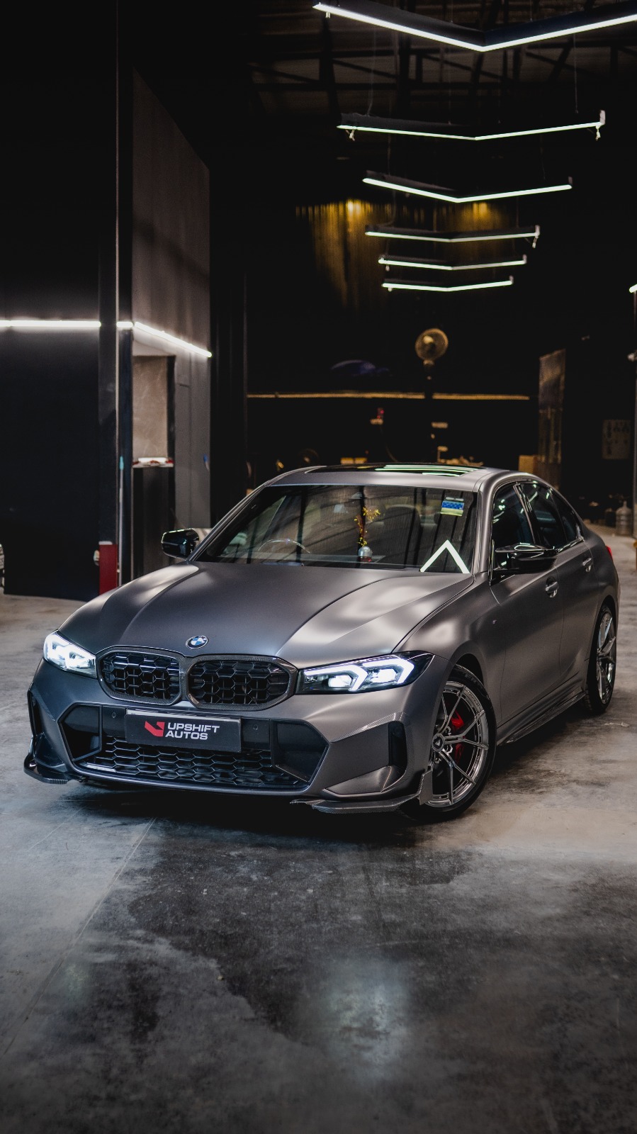 A BMW 3 Series in satin grey with a black kidney grille, front three-quarter view on the workshop floor.