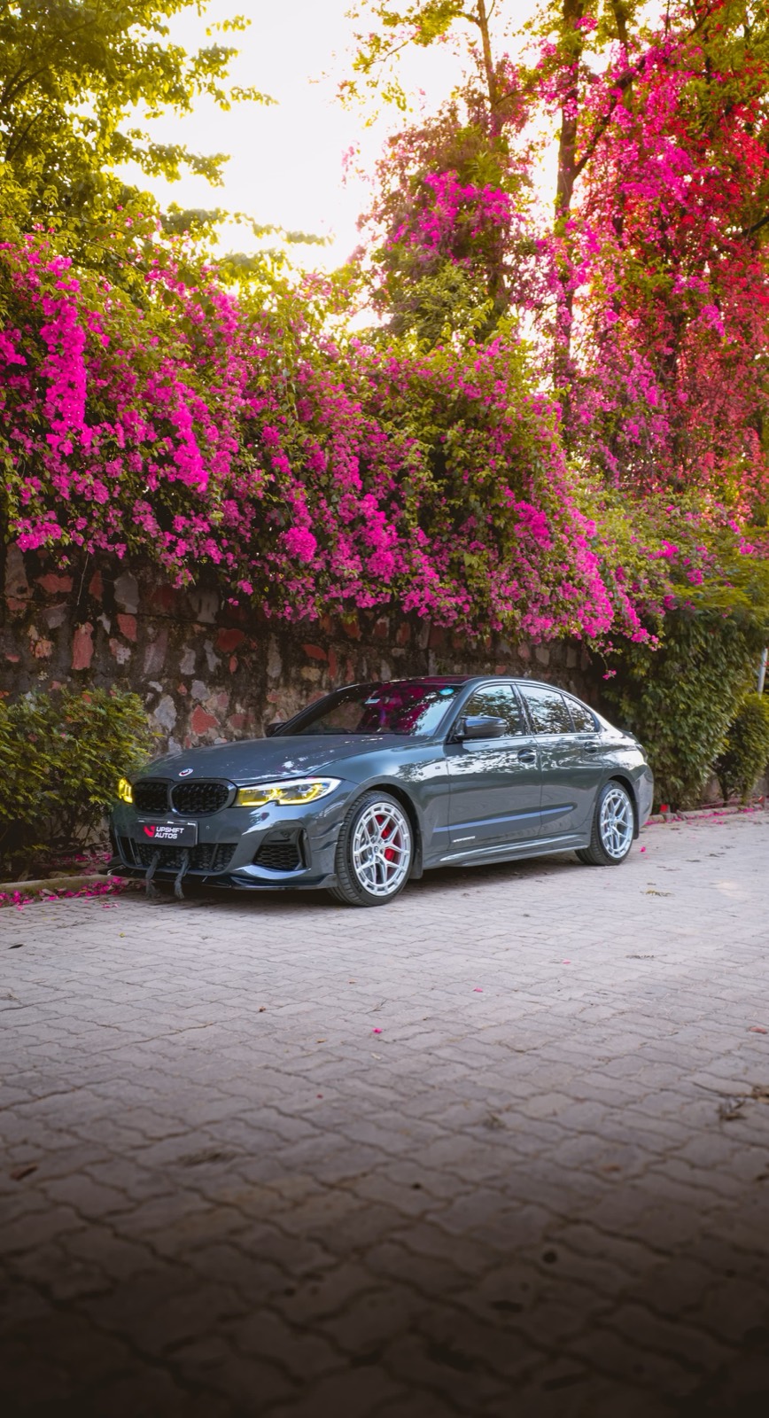 A grey BMW saloon with yellow headlamps and silver wheels, front three-quarter view, parked on a paved lane beneath a stone wall covered in pink bougainvillea.