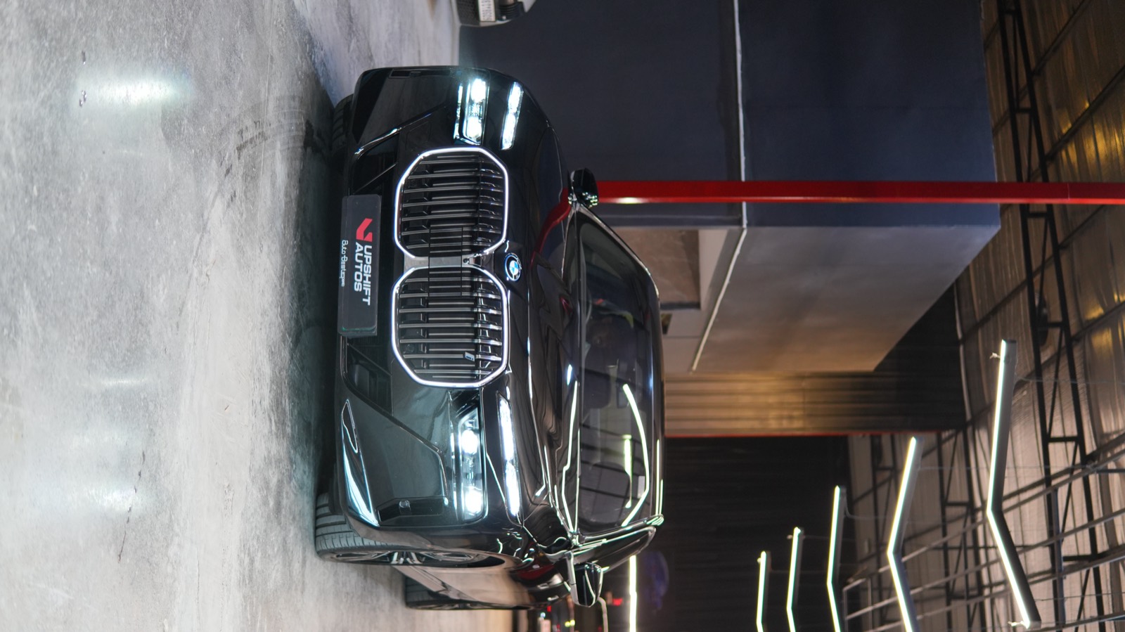 A black BMW i7 with its illuminated kidney grille and headlamps lit, head-on in the workshop hall beneath angled strip lights and a red steel column.