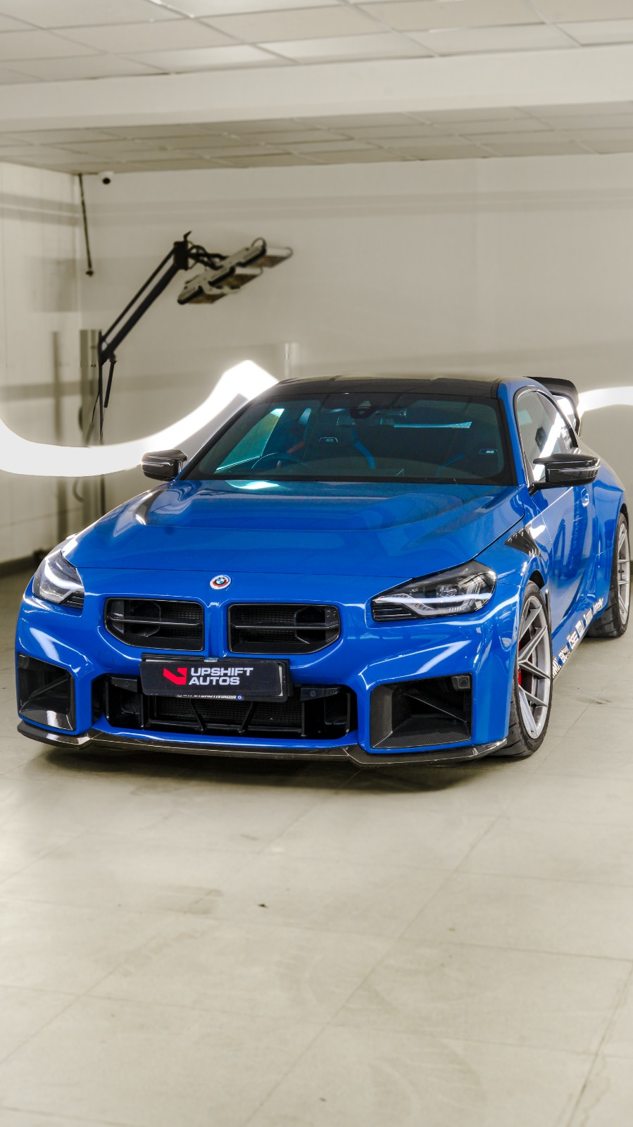 A bright blue BMW M2 with a carbon roof and rear wing, front view in a white-walled bay.