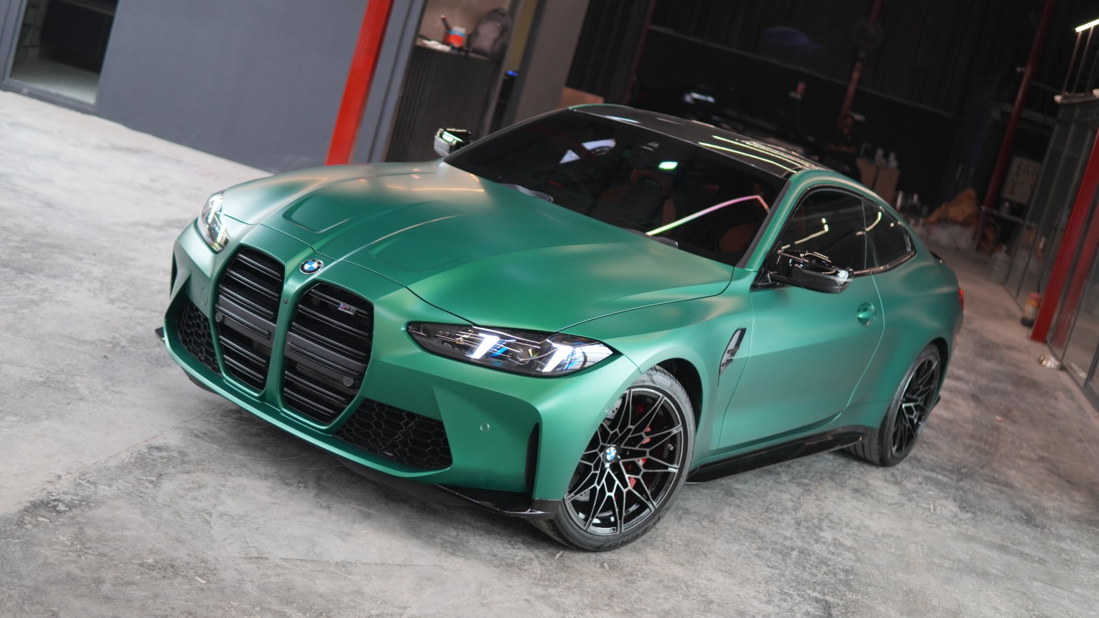 A green BMW M4 coupé in a satin finish, front three-quarter view in the workshop with black multi-spoke wheels.