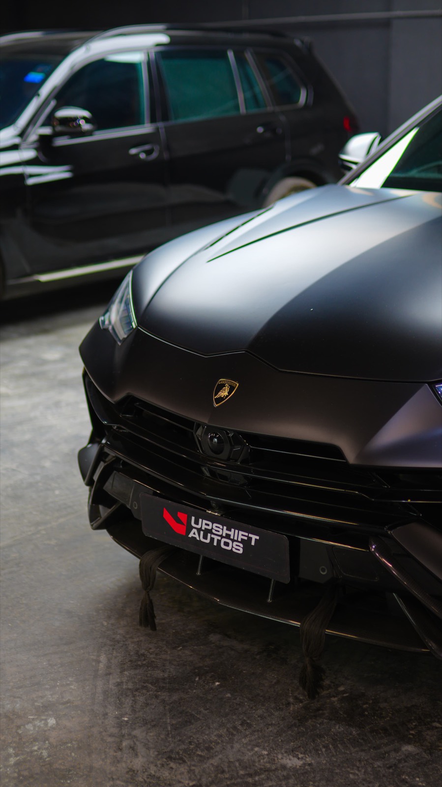 Close view of the matte black nose of a Lamborghini Urus in the workshop, with a gloss black SUV parked behind it.
