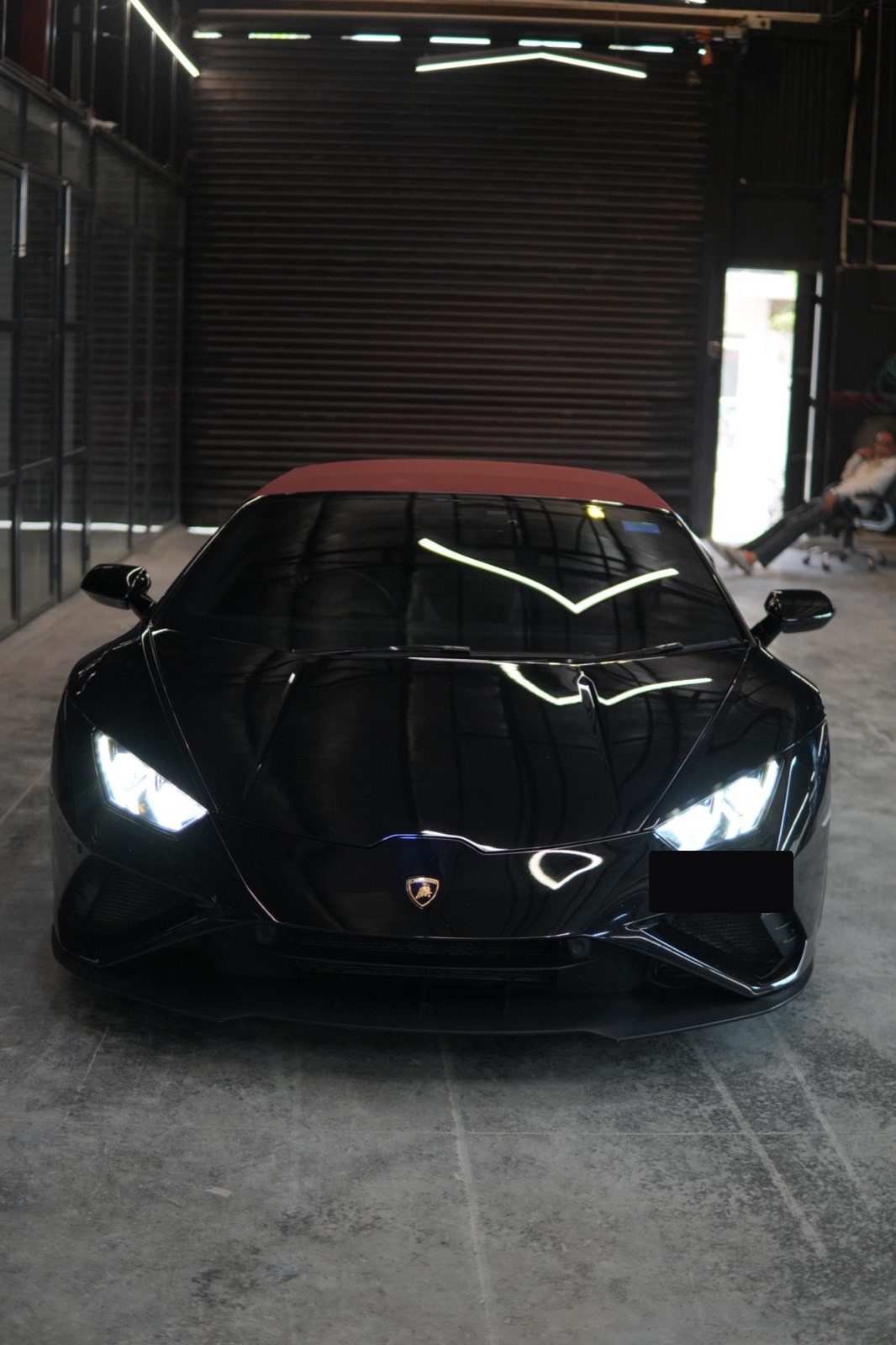 A black Lamborghini Huracán convertible with a red soft top, headlamps lit, facing the camera head-on inside the workshop hall.