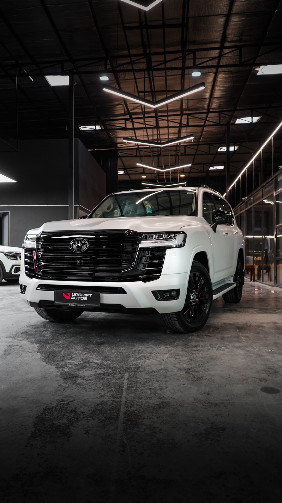 A white Toyota Land Cruiser LC300 in a satin finish with a black grille and black wheels, front three-quarter view in the workshop hall.