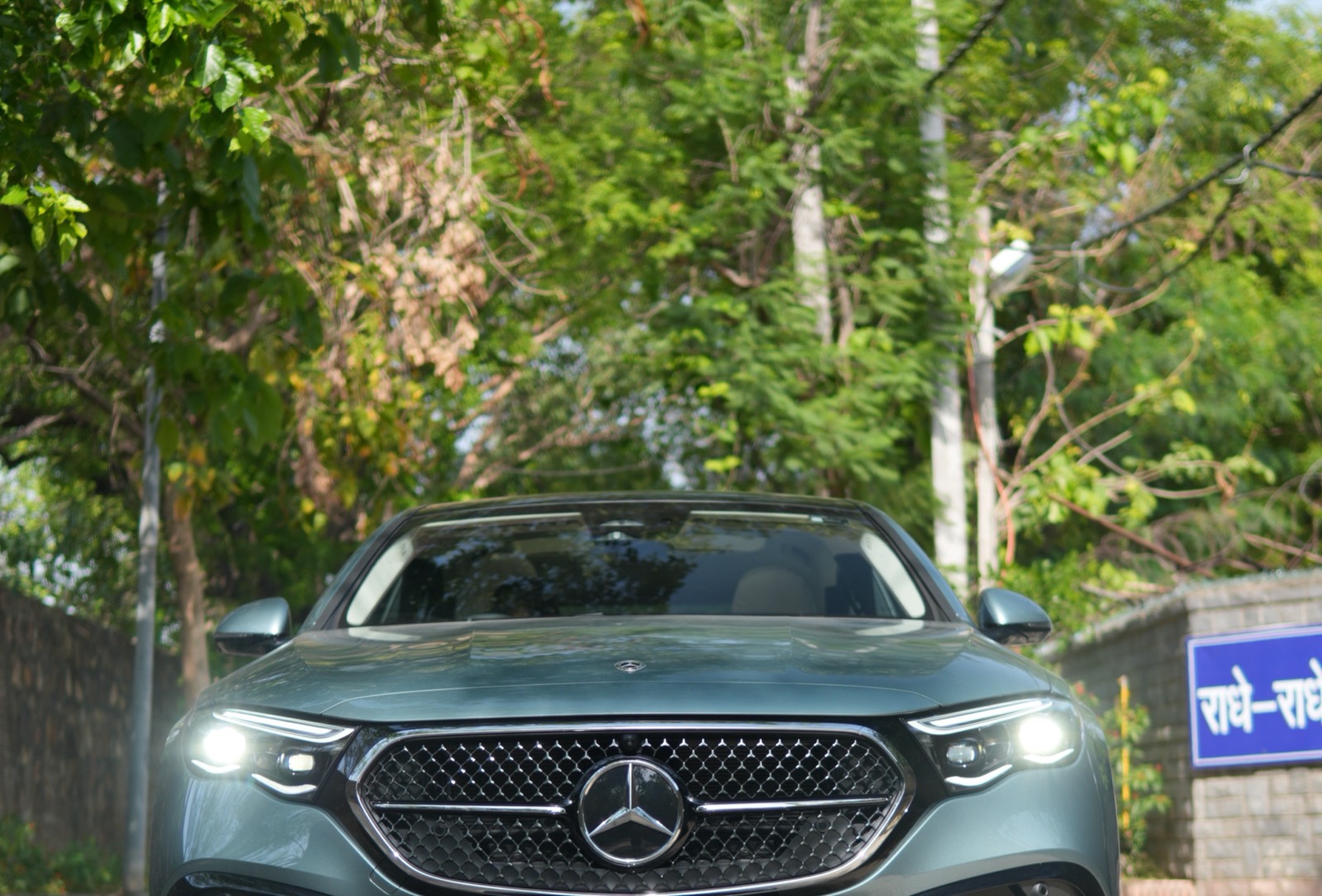 A grey-green Mercedes-Benz E-Class, head-on with its headlamps lit, on a tree-lined lane outdoors.