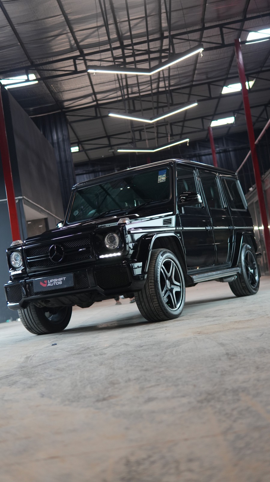 A black Mercedes-AMG G63, front three-quarter view in the workshop hall beneath chevron strip lights.