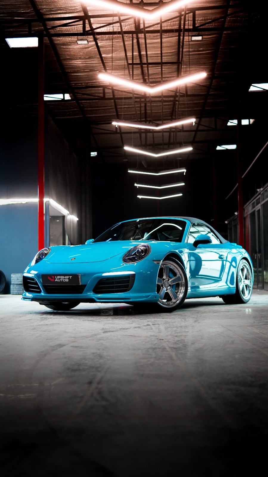 A bright blue Porsche 911 Cabriolet with its black roof up, front three-quarter view in the workshop hall.