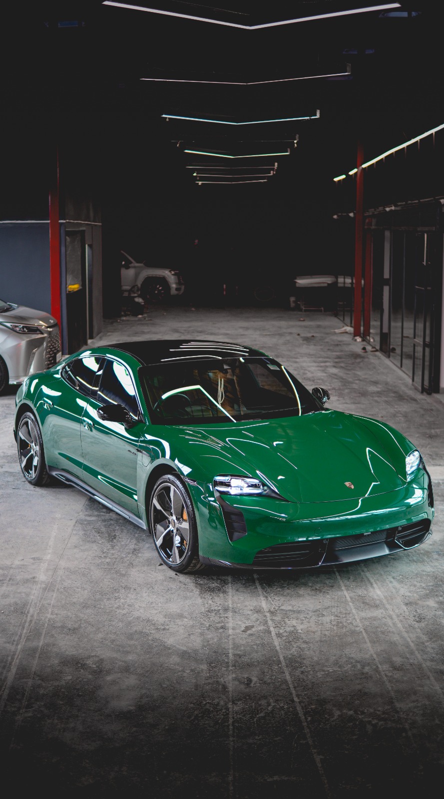 A dark green Porsche Taycan, front three-quarter view in the workshop hall under red steel columns.