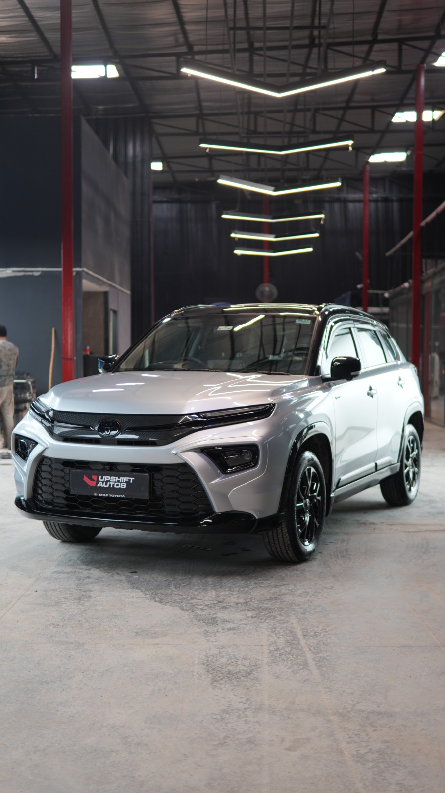 A silver Toyota Urban Cruiser Hyryder with a black roof, front three-quarter view in the workshop hall.
