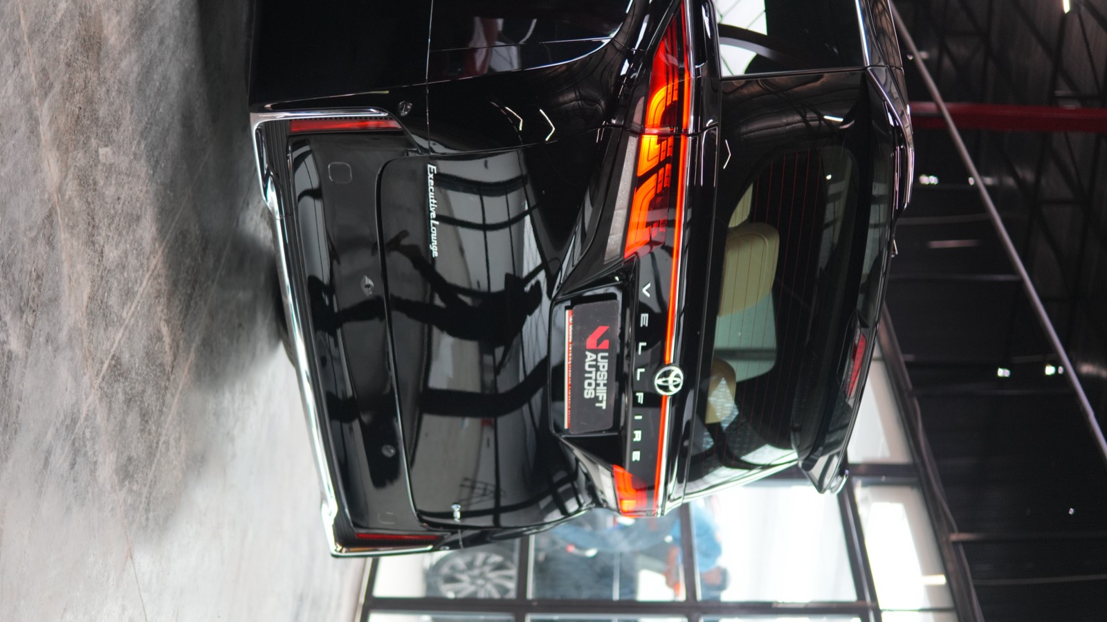 The rear of a black Toyota Vellfire Executive Lounge in the workshop, tail lamps lit, with a figure reflected in the gloss of the tailgate.